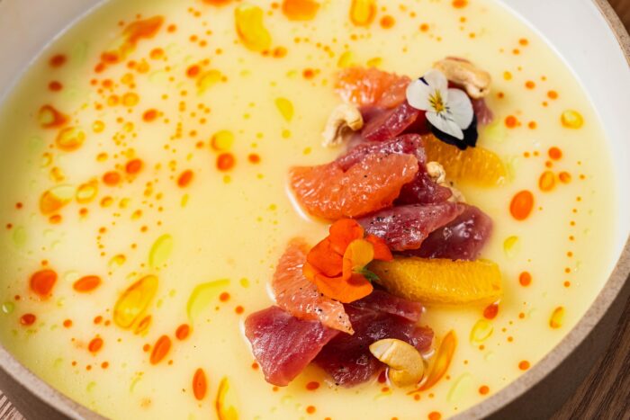 A creamy yellow soup bowl from Olive and Fig, garnished with raw tuna, citrus segments, cashew nuts, and edible flowers.