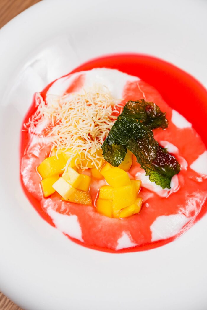 A bowl of white yogurt topped with diced mango, shredded coconut, an olive leaf, and a fig puree drizzle.