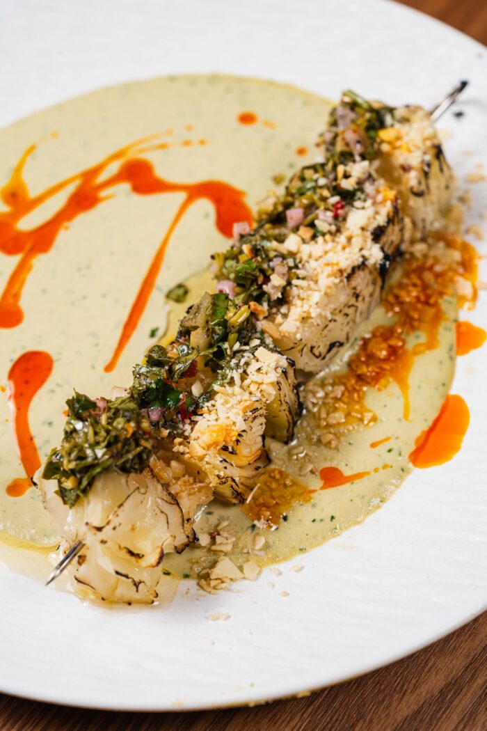 A skewer of charred olive and fig vegetable segments topped with herbs and crushed nuts, served over a green sauce drizzled with chili oil.