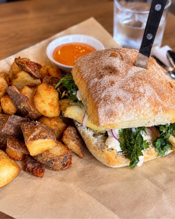 An Olive and Fig ciabatta sandwich with a knife stuck in the top, served with a side of roasted potatoes and a small cup of orange dipping sauce.