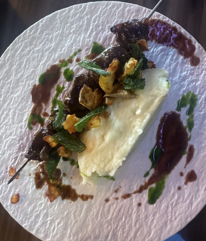 A meat skewer with olive and fig garnishes served over mashed potatoes with drizzles of dark and green sauce on a white plate.