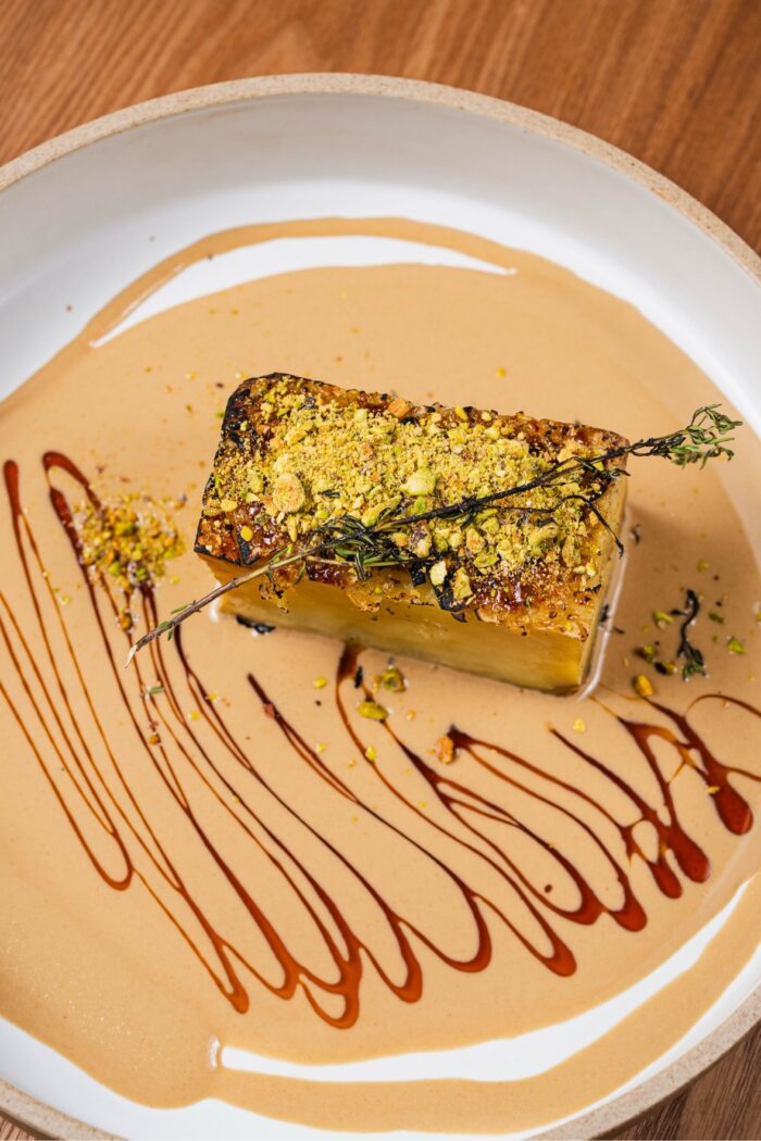 A square serving of olive and fig potato gratin topped with crushed pistachios and a sprig of thyme, drizzled with sauce on a plate.