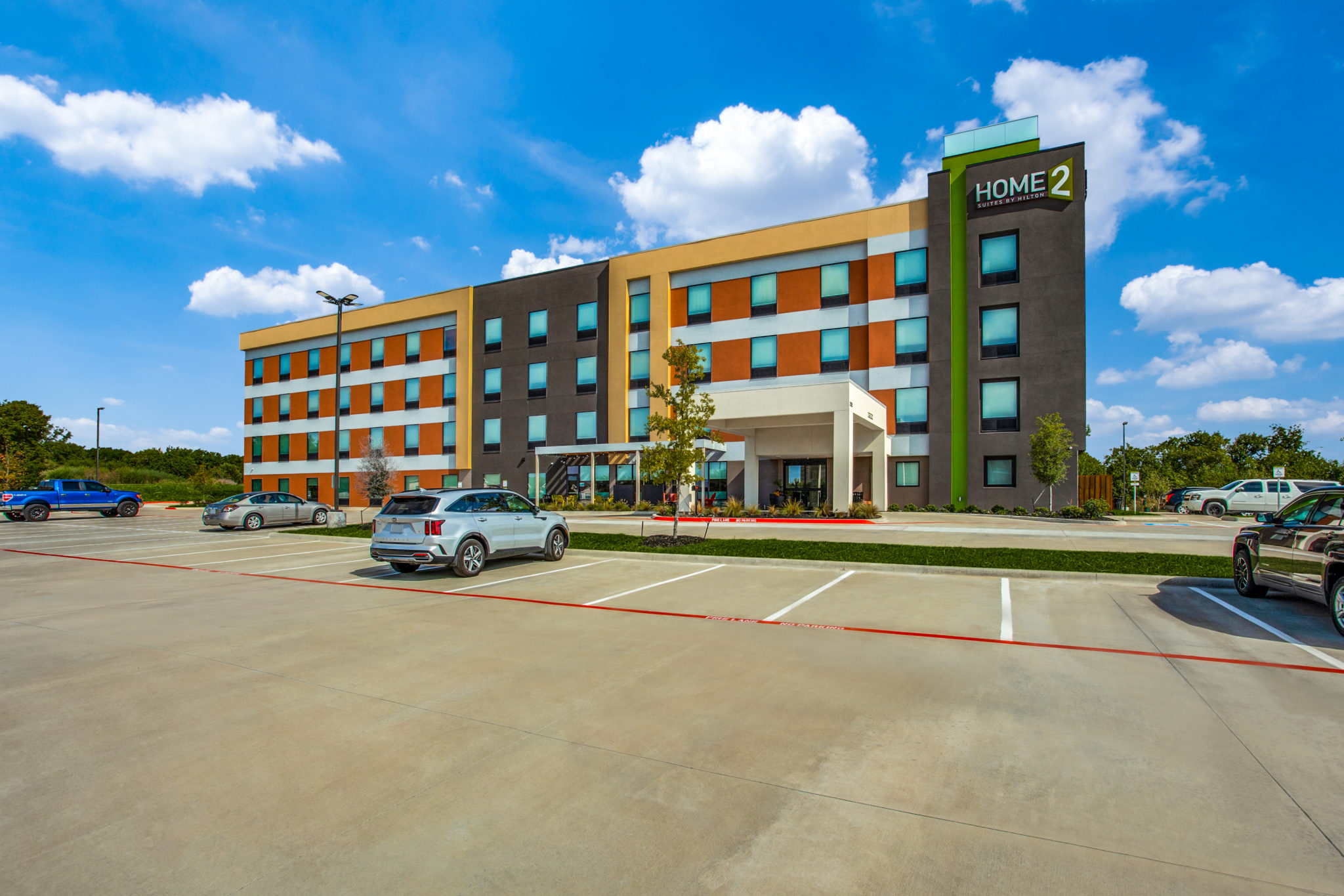 A modern, four-story Home2 Suites hotel with colorful exterior panels offers exceptional hotel deals, welcoming guests to enjoy several parked cars in front, landscaped greenery, and a clear blue sky with scattered clouds.