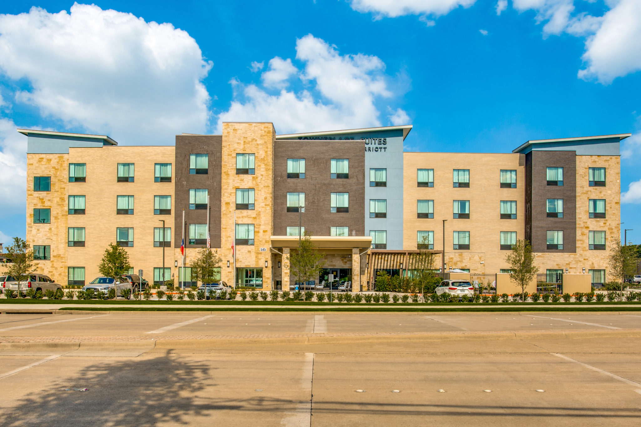 A modern four-story hotel building with beige and dark gray exterior panels, large windows, and a covered entrance. Several cars are parked in front under a bright sky—perfect for travelers searching for great hotel deals.