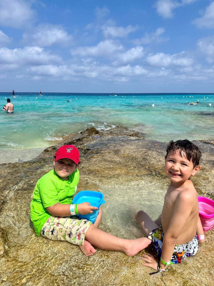 Two young boys sit on rocky shore in shallow water, smiling. One wears a red cap and green shirt, holding a blue bucket; the other is shirtless in swim trunks. Bright blue ocean and swimmers are in the background under a partly cloudy sky.