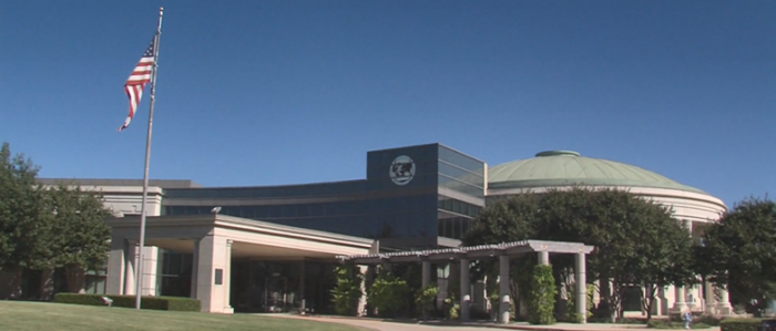 Modern building with a domed roof, an American flag on a pole, green lawn, and trees surrounding the entrance under a clear blue sky.