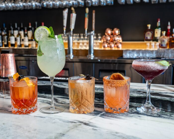 Five colorful cocktails on a marble bar counter, each garnished differently with fruit or herbs, with bottles and glasses in the background.