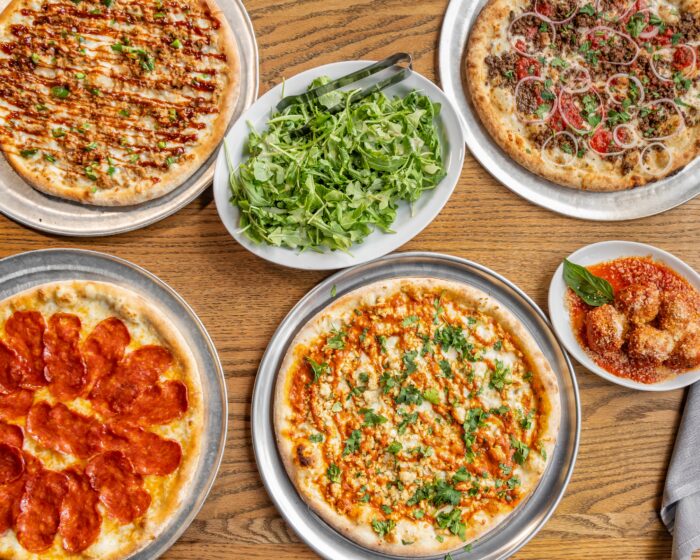 Four pizzas with different toppings, a plate of arugula salad with tongs, and a small bowl of arancini with marinara sauce and a basil leaf are arranged on a wooden table.
