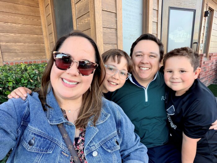 Four people, two adults and two children, smile for a selfie outside a wooden building on a sunny day. The woman in front wears sunglasses and a denim jacket; the others are close together, looking happy.