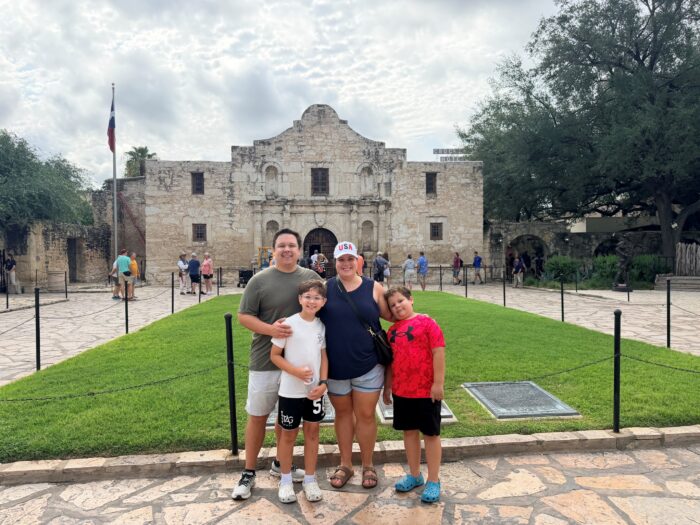 A family of four poses, smiling in front of the historic Alamo Mission in San Antonio, Texas, on a cloudy day. Other visitors and lush green grass are visible in the background.