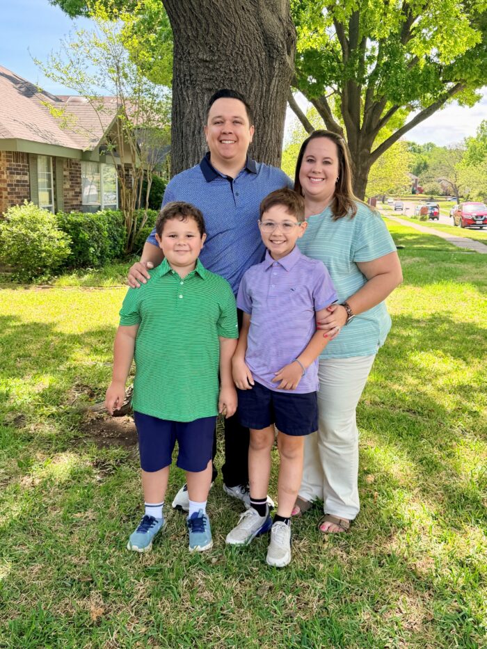 A smiling family of four stands together on green grass under a large tree, with houses and parked cars visible in the background on a sunny day.