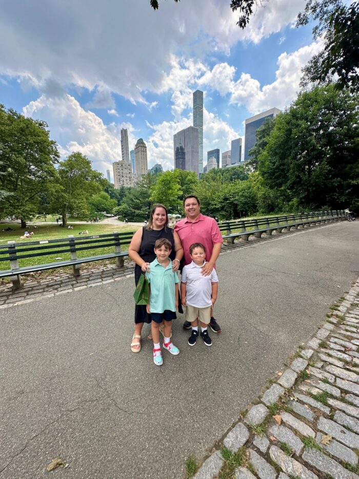 A family of four poses on a park pathway with tall city buildings and trees in the background under a partly cloudy sky.