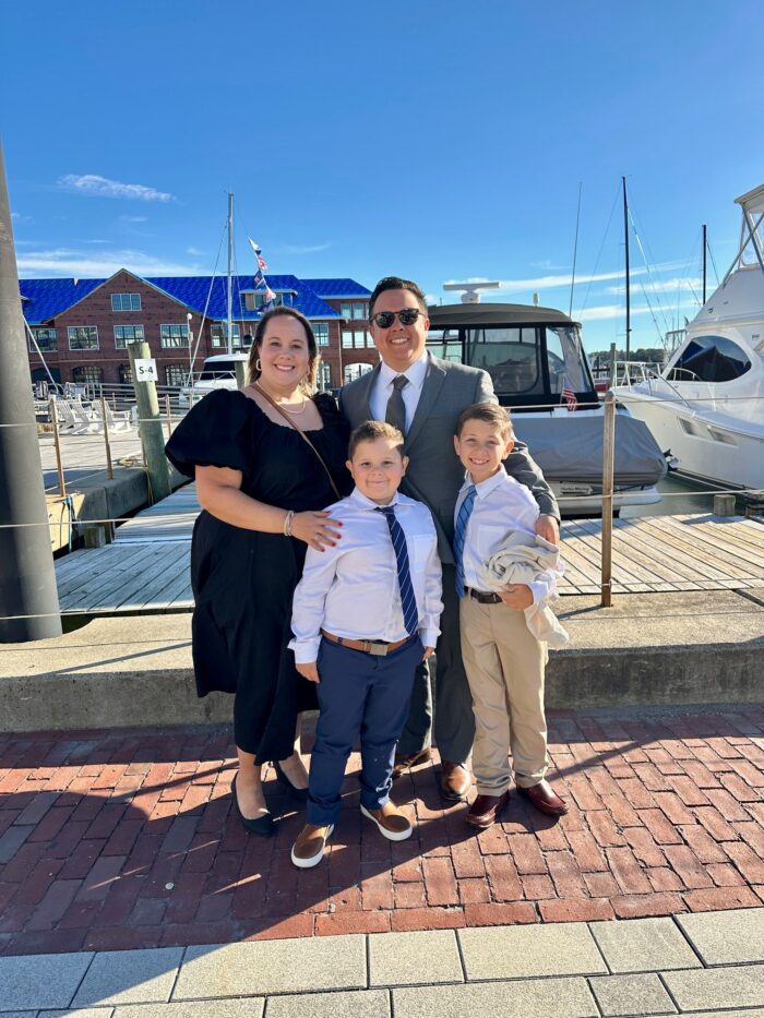 A family of four stands smiling on a marina dock. The parents are dressed formally, and their two young sons wear dress shirts and ties. Boats and a brick building with a blue roof are in the background under a clear blue sky.