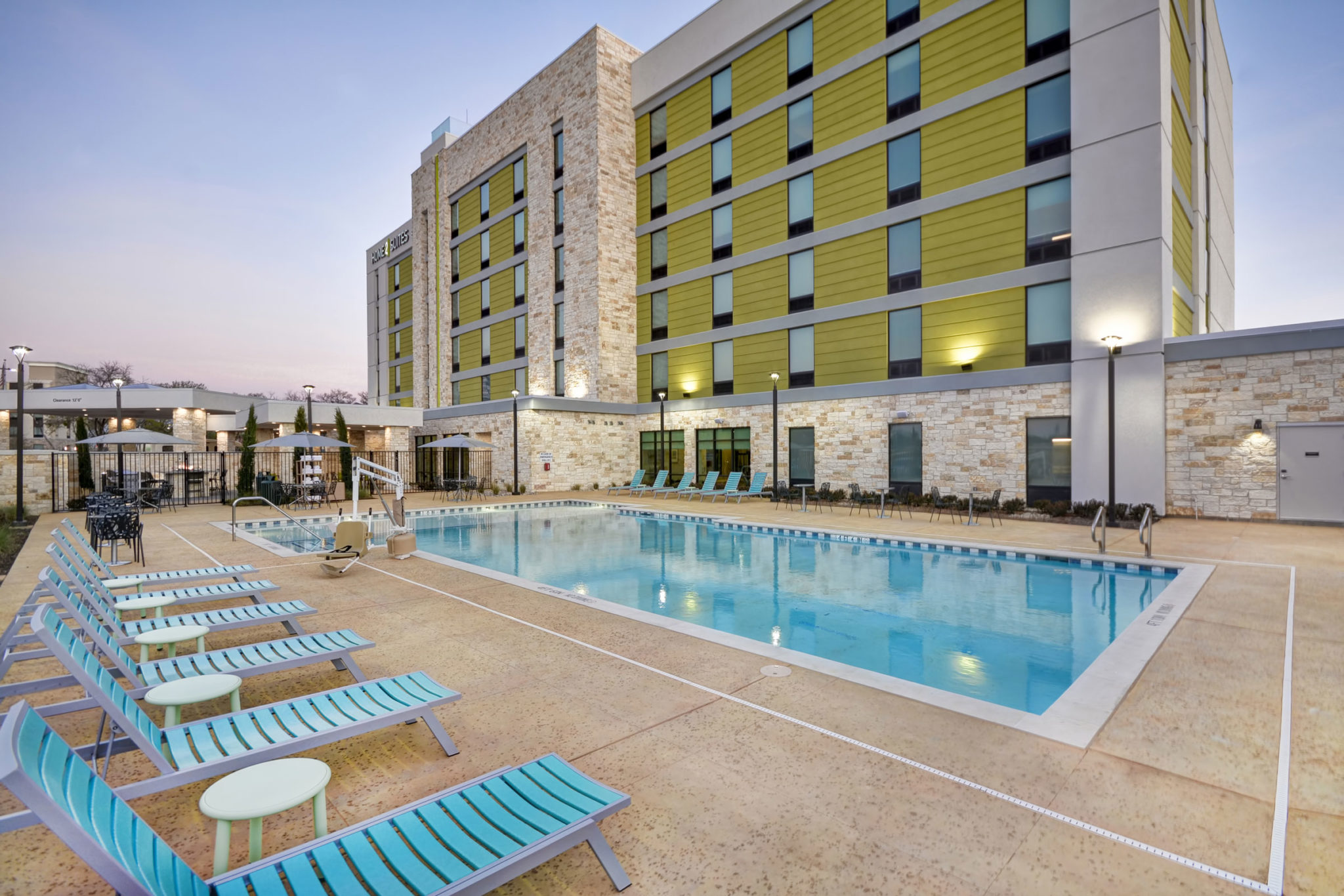 Outdoor hotel pool with turquoise lounge chairs lined up on the deck. Behind the pool is a modern, multi-story building with green and beige accents. Enjoy this clean, well-lit retreat in the early evening—ask about exclusive hotel deals today.