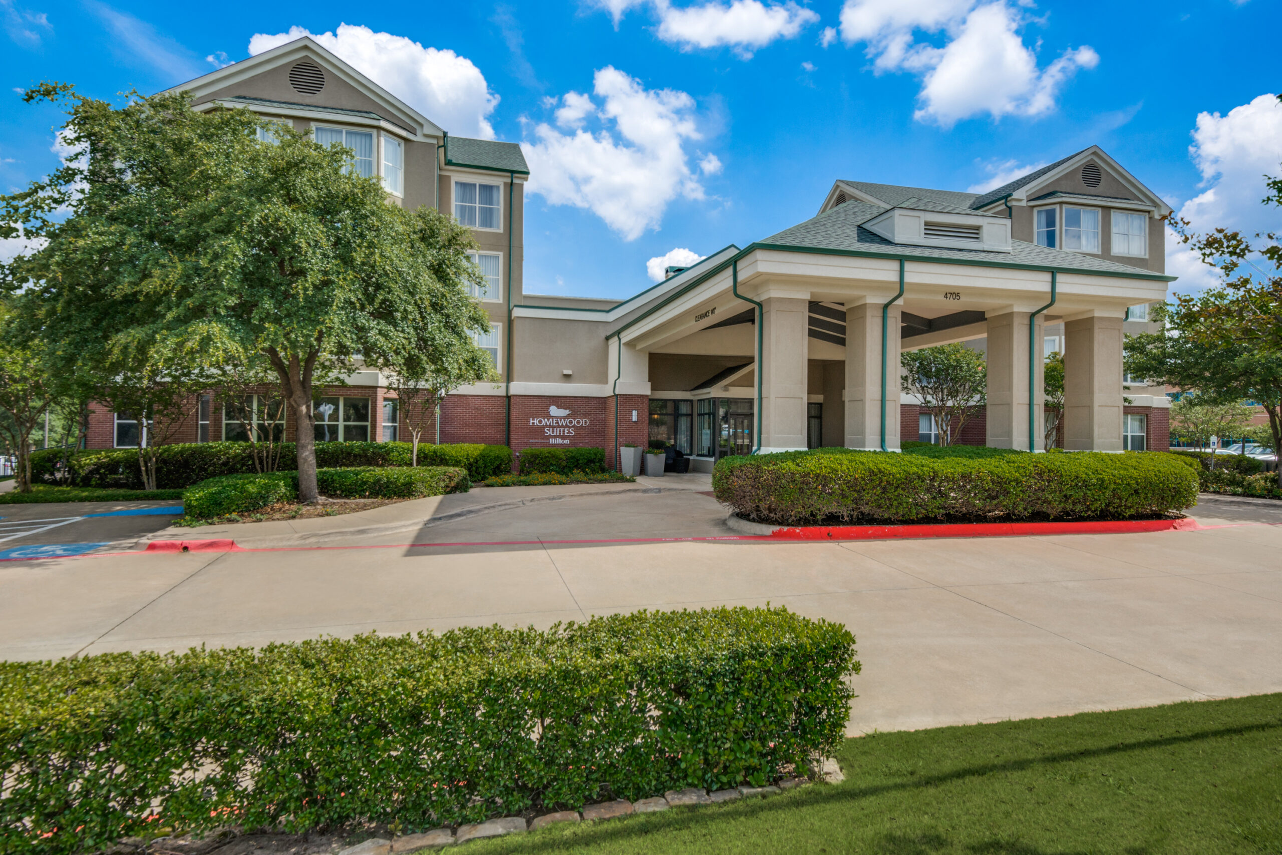 A modern hotel building with beige walls and green roofing, surrounded by trimmed shrubs and trees, offers inviting hotel deals. A driveway winds under a clear blue sky dotted with clouds overhead.