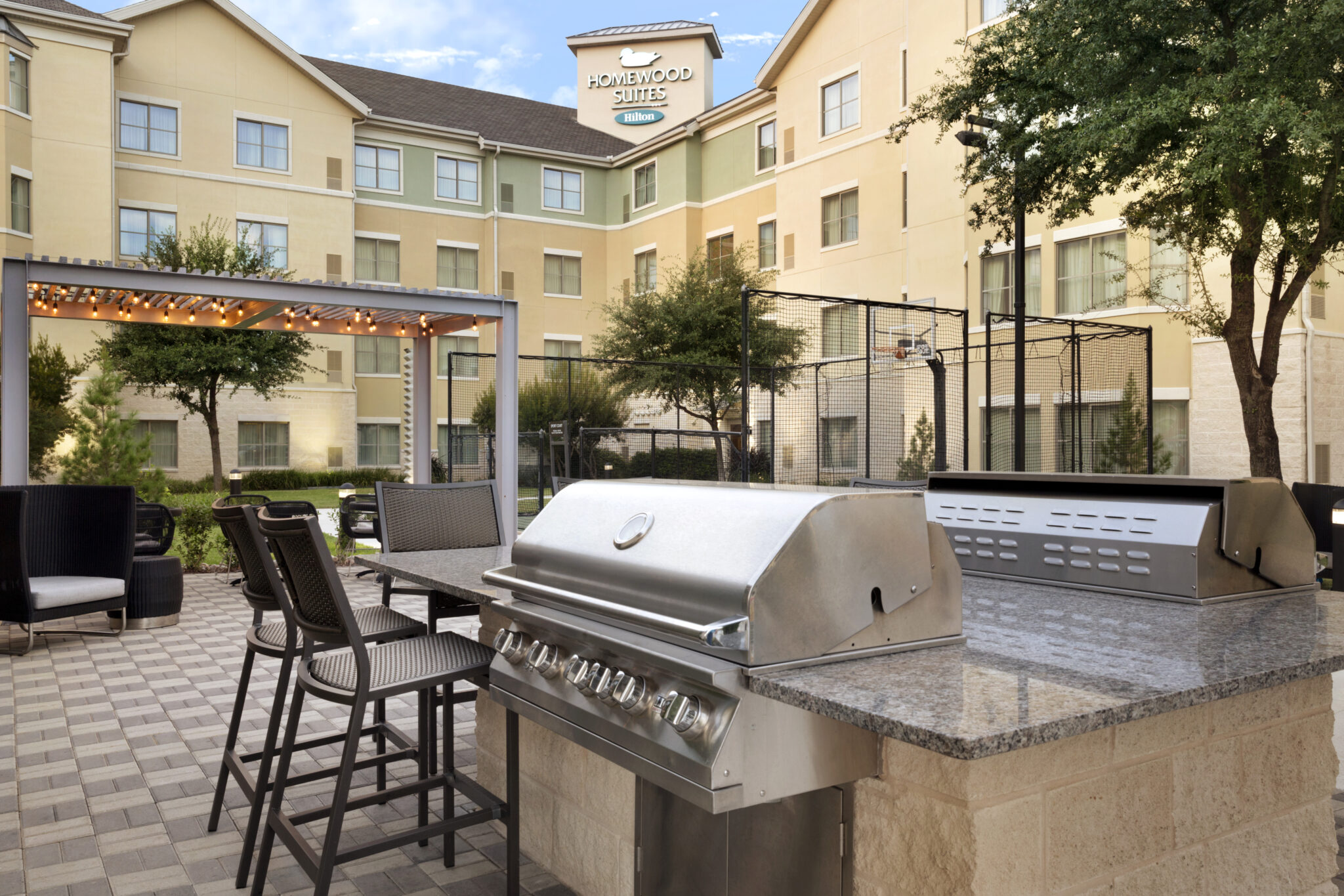 An outdoor patio area at Homewood Suites featuring a stainless steel grill, bar seating, lounge chairs, a pergola with string lights, and landscaped greenery—perfect for relaxing after exploring the latest hotel deals.