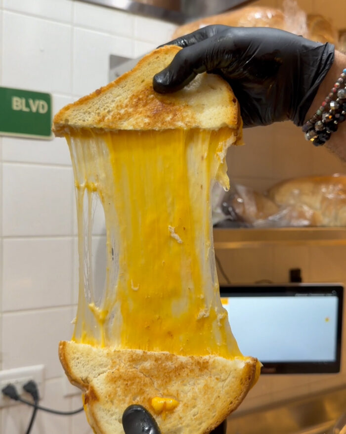 A gloved hand pulls apart a grilled cheese sandwich, showing long, stretchy melted cheese between two toasted bread slices in a kitchen setting.