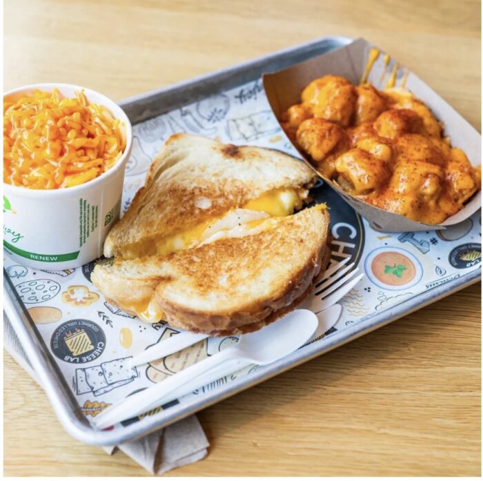 A tray with a grilled cheese sandwich, a paper cup of macaroni and cheese, and a paper dish of saucy tater tots, with a white plastic fork and spoon on a colorful paper liner.
