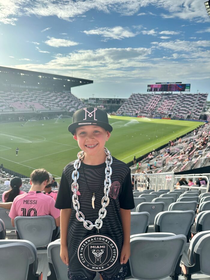 A smiling child wearing an Inter Miami jersey, large chain necklace, and black cap stands in a stadium with a soccer field and fans in the background.
