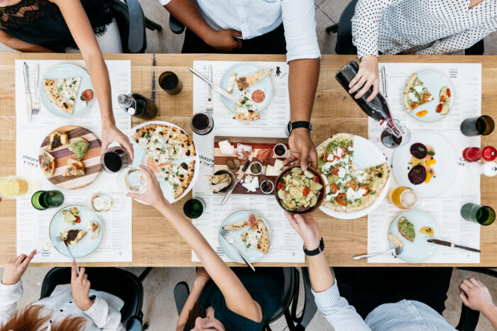 A group of people share pizza, drinks, and appetizers at a wooden table, reaching for food and drinks, seen from above. Plates, glasses, and menus are arranged neatly around the table.