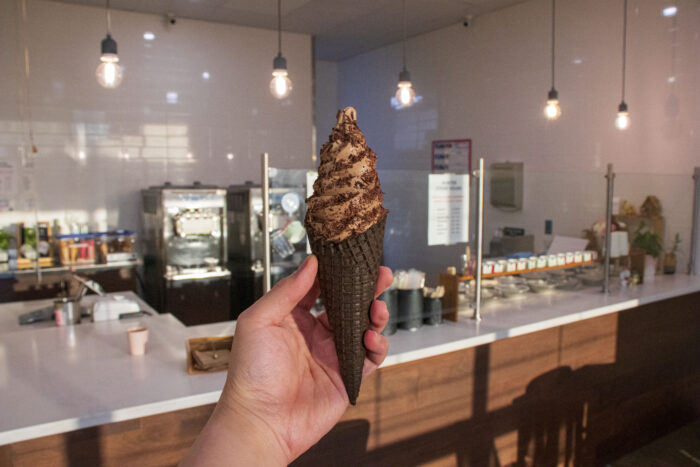 A hand holds a chocolate soft serve ice cream in a dark waffle cone inside a modern, well-lit ice cream shop with hanging lights and a white countertop, perfectly capturing the joy of enjoying fresh ice cream.