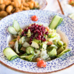 A bowl of fresh salad with lettuce, cucumber, chopped vegetables, pomegranate seeds, and cherry tomatoes, served in a decorative blue and white dish. Fried appetizers and other plates are visible in the background.