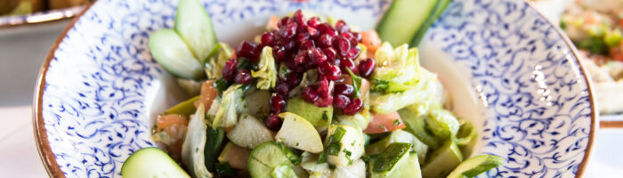 A bowl of fresh salad with lettuce, cucumber, chopped vegetables, pomegranate seeds, and cherry tomatoes, served in a decorative blue and white dish. Fried appetizers and other plates are visible in the background.