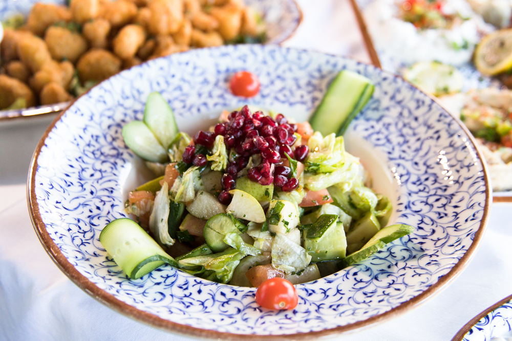 A bowl of fresh salad with lettuce, cucumber, chopped vegetables, pomegranate seeds, and cherry tomatoes, served in a decorative blue and white dish. Fried appetizers and other plates are visible in the background.