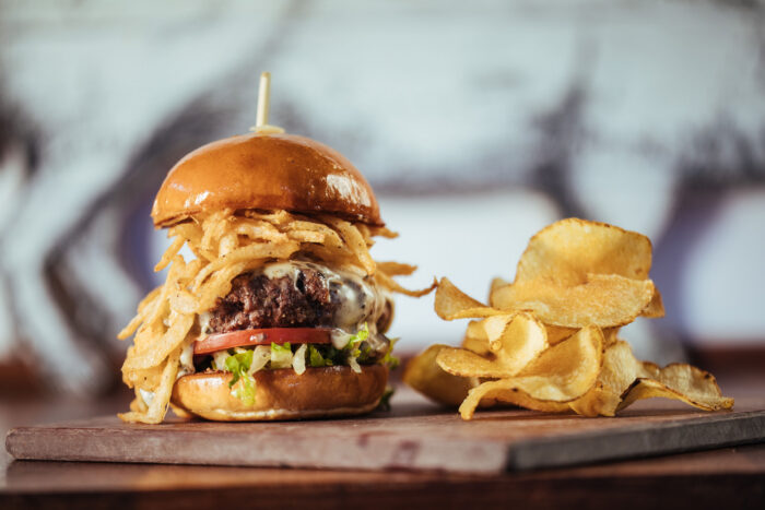 A cheeseburger with lettuce, tomato, and crispy fried onions on a wooden board, served with a side of potato chips.
