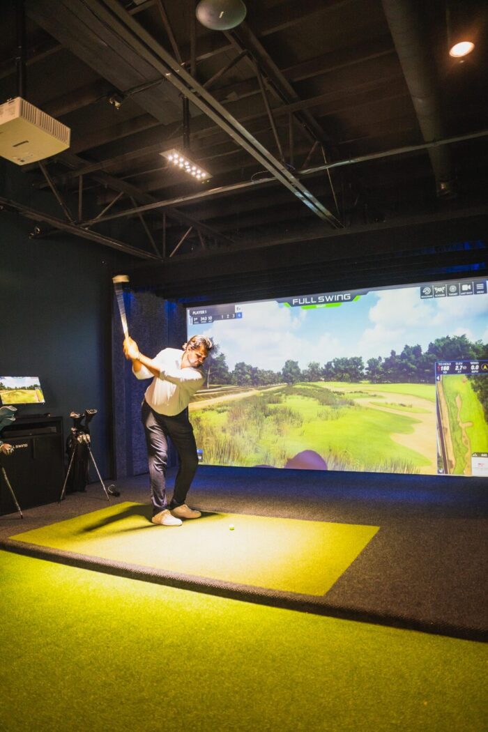 A person swings a golf club on an indoor golf simulator, aiming at a virtual golf course displayed on a large screen.