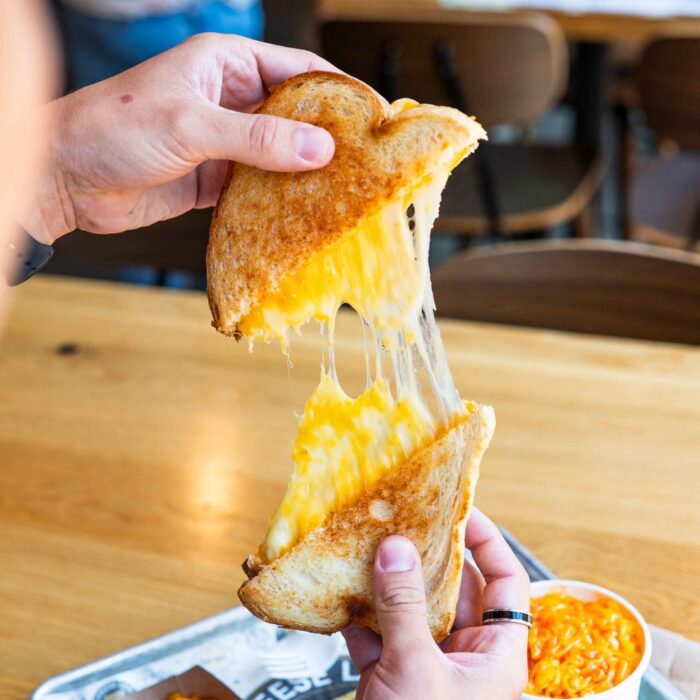 A person pulls apart a grilled cheese sandwich, showing gooey melted cheese stretching between the toasted bread halves. A bowl of shredded cheese and a tray are on the wooden table below.