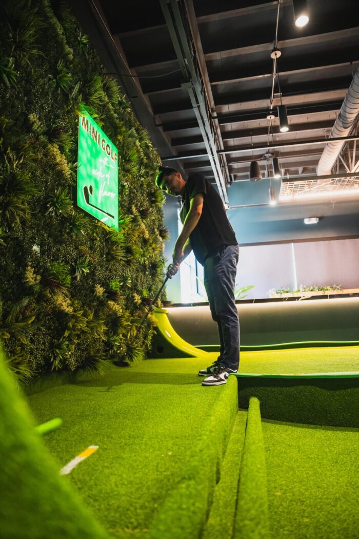 A person playing mini-golf indoors, standing on artificial turf with a putter, next to a green wall and a neon sign that reads Minigolf..