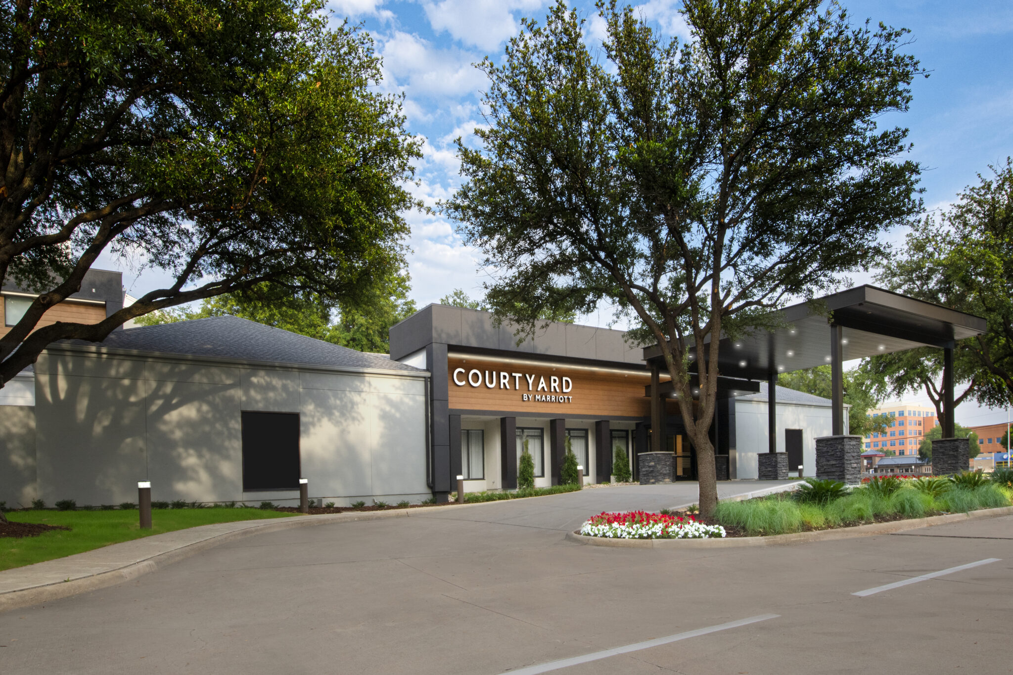 A modern Courtyard by Marriott hotel entrance with a covered driveway, surrounded by trees, greenery, and flowers under a blue sky—discover exclusive hotel deals for your next stay.