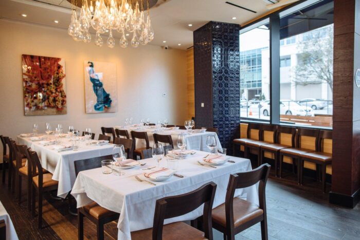 Elegant restaurant dining area with white tablecloths, wooden chairs, and modern art on the walls. Large windows provide natural light, and a chandelier hangs from the ceiling. Tables are set with glasses and napkins.