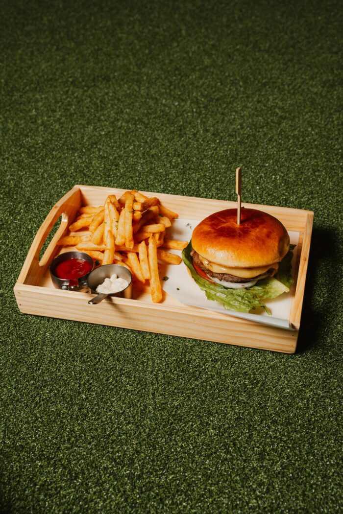 A wooden tray with a hamburger, a serving of French fries, ketchup, and mayonnaise sits on green artificial grass.