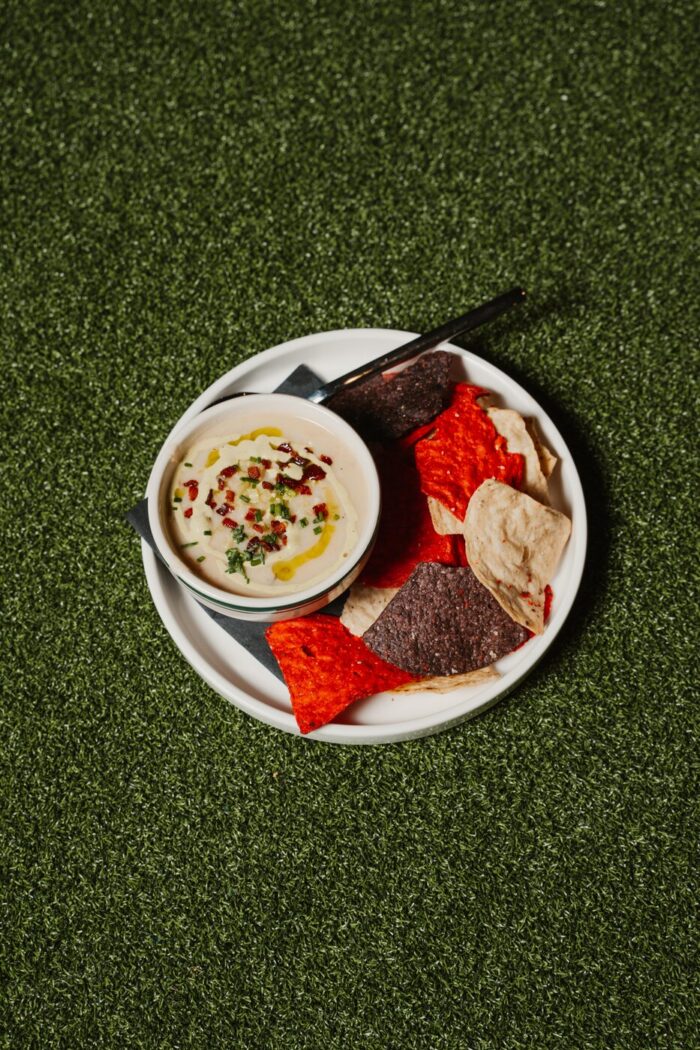A bowl of creamy dip garnished with herbs and oil, served with assorted colorful chips on a white plate, placed on green artificial grass.