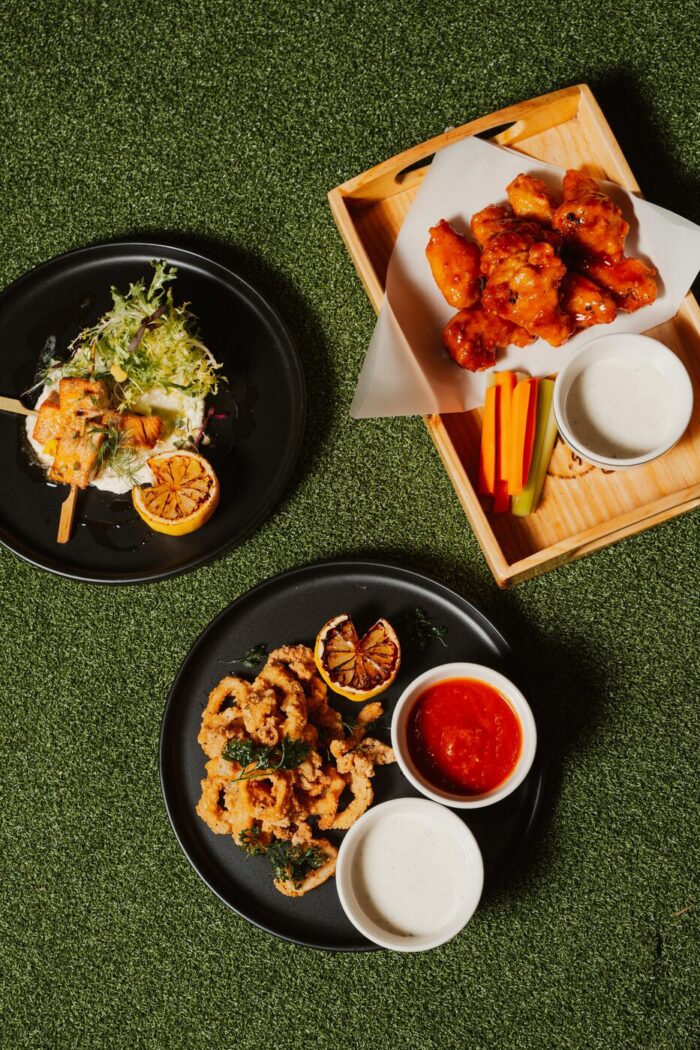 Three plates of food on green grass: grilled skewers with salad, saucy chicken wings with celery and carrot sticks, and fried calamari with dipping sauces and grilled lemon.
