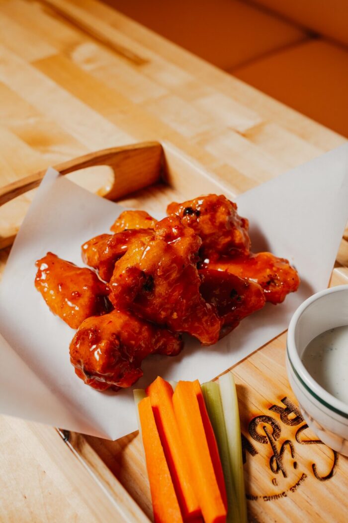 A serving of saucy buffalo chicken wings on parchment paper with carrot and celery sticks and a cup of ranch dip on a wooden tray.
