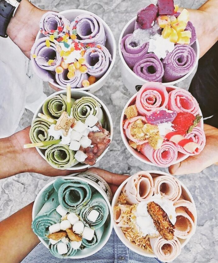 Five hands hold colorful ice cream cups filled with rolled ice cream, each topped with fruit, chocolate, marshmallows, and sprinkles, arranged in a cheerful circle on a gray surface.