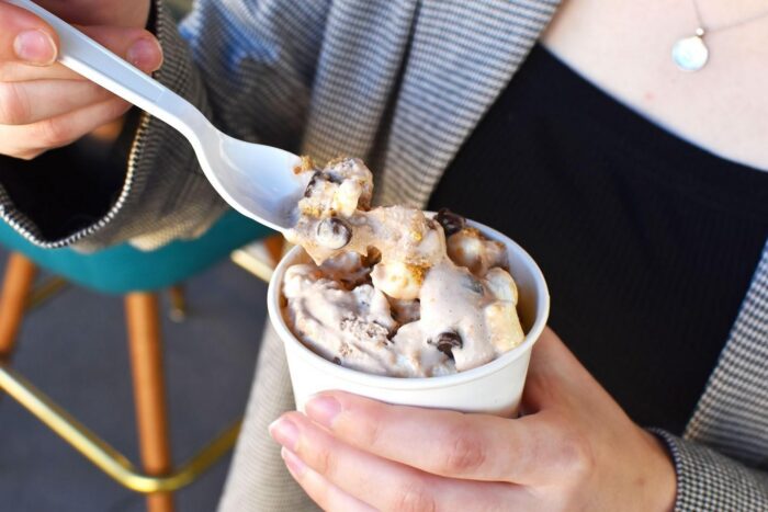 A person holding a cup of ice cream topped with chocolate chips and marshmallows, scooping some with a plastic spoon. The person is wearing a striped jacket and a necklace, enjoying their delicious ice cream treat.