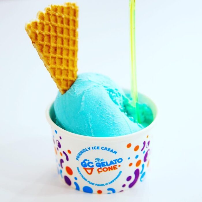 A cup of bright blue gelato ice cream with a triangular waffle piece and a translucent green spoon, served in a white cup with colorful dots and The CC Gelato Cone logo on it.
