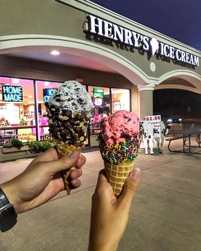 Two hands hold up ice cream cones outside Henry’s Ice Cream shop at night. One cone has cookies and cream with chocolate chips; the other, pink ice cream with rainbow sprinkles. The shop glows with colorful signs, capturing the joy of an ice cream outing.
