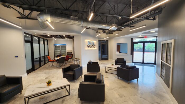 Modern office lobby with marble floors, black leather chairs, small tables with plants, glass-walled meeting room, industrial ceiling, and a sign on the wall reading SUITE WORK near large windows and a glass door.