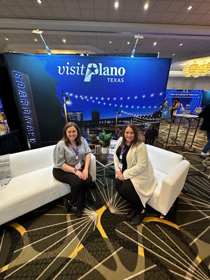 Two women sit on white couches in front of a Visit Plano Texas display booth at an indoor event, utilizing Visit Plano brochures and decor arranged on a small table between them.
