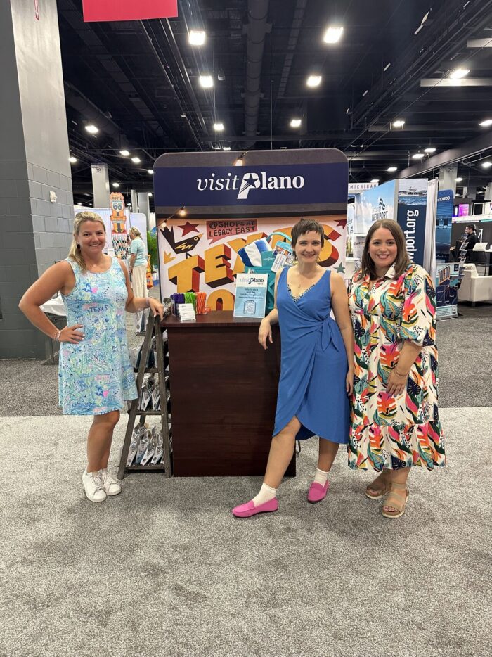 Three women in colorful dresses stand smiling in front of a Visit Plano booth at a convention or expo. The booth displays brochures and promotional materials. The setting is bright and spacious.
