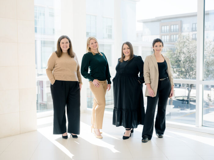 Four women stand in a bright, modern space with large windows. They wear business-casual outfits and smile at the camera. Sunlight streams in, creating a light and airy atmosphere—an inspiring setting that highlights why so many professionals choose to utilize Visit Plano for meetings and events.