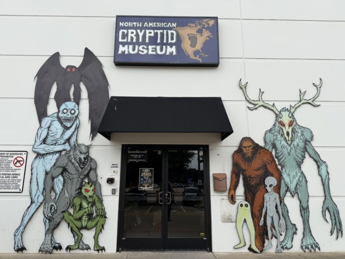 Entrance to the North American Cryptid Museum with large cutouts of various cryptids, including Bigfoot, Mothman, and other mythical creatures, displayed on either side of the door.