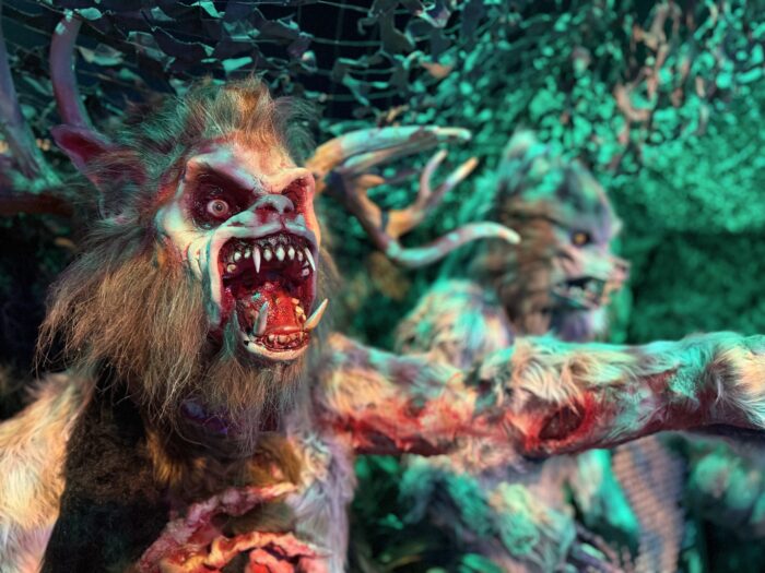 Two snarling, monstrous creatures with sharp teeth and claws, covered in matted fur and blood, stand under greenish lighting and camouflage netting, creating a frightening and eerie atmosphere.