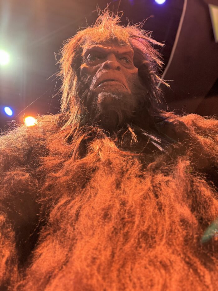 A low-angle shot of a tall, hairy creature with reddish fur and a serious expression, under dramatic lighting with orange and yellow highlights.