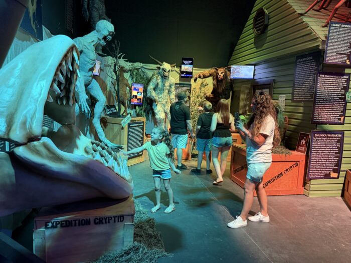People look at cryptid monster exhibits and read plaques in a museum-style setting, with large creature sculptures and informational displays under blue and yellow lighting.